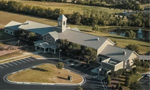 Aerial view of a large church complex with a central tower, surrounded by parking lots, trees, and open green spaces. There is a pond with fountains in the background.